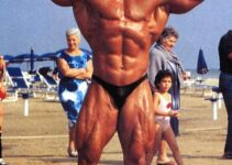 Mike Quinn doing a shirtless front double biceps flex on the beach, looking huge and ripped