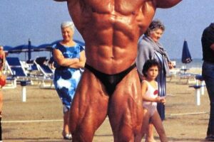 Mike Quinn doing a shirtless front double biceps flex on the beach, looking huge and ripped