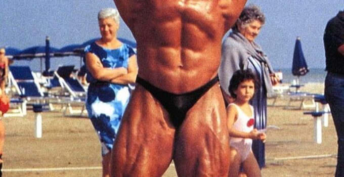 Mike Quinn doing a shirtless front double biceps flex on the beach, looking huge and ripped