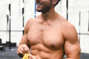 Khan Porter eating a snack while being shirtless in a CrossFit gym, looking ripped and strong