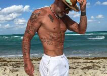 Gianluca Vacchi stainding on the beach with a hat on, looking fit